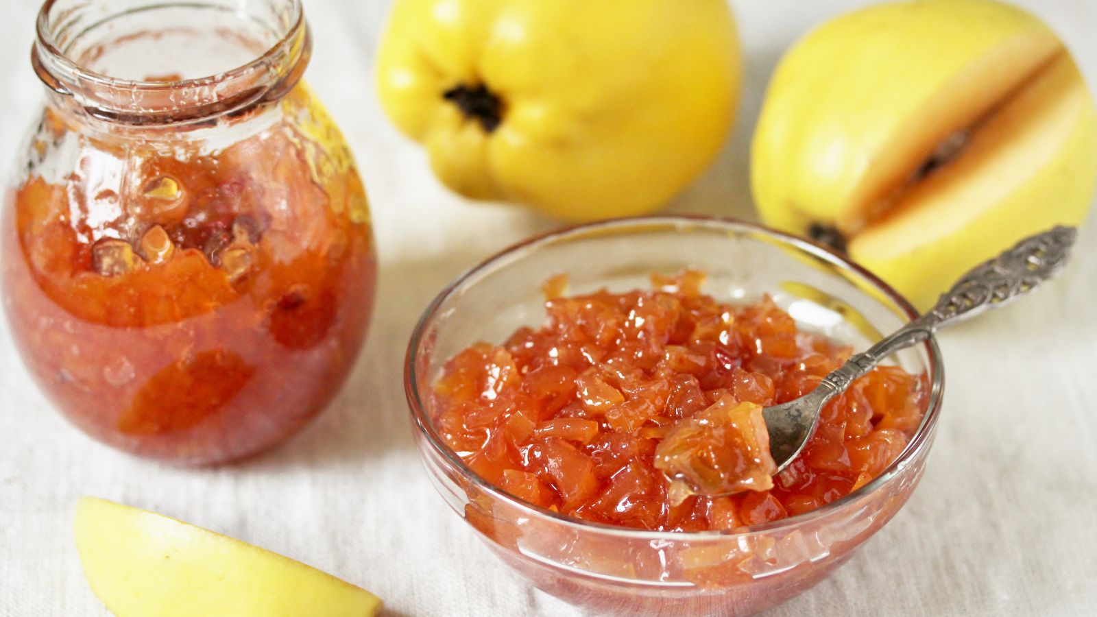 A shot of a fruit jam in a jar and a glass bowl made from the fresh pear-shaped fruit in the background that are all placed on top of a surface with a table cloth