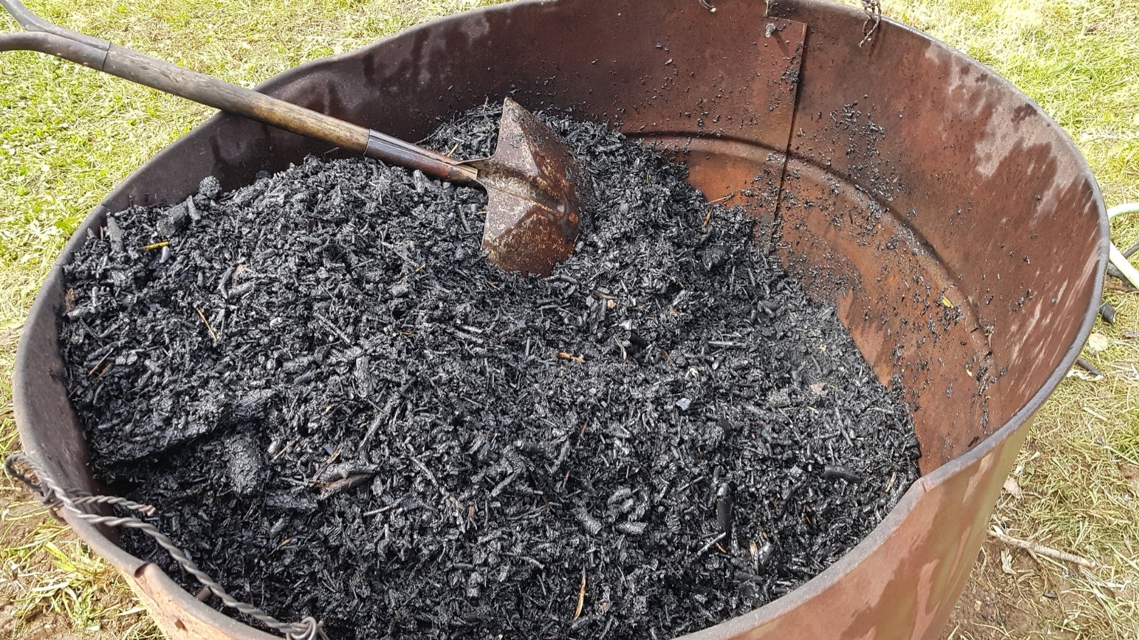 A bucket filled with dark, charred biochar sits on the ground with a wooden spatula resting inside, its blackened, porous texture contrasting against the garden backdrop.