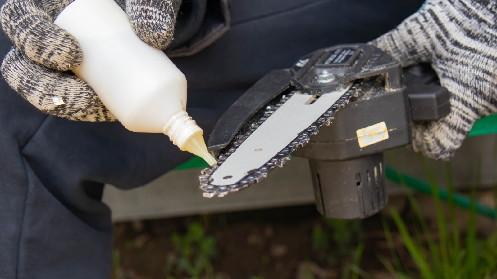 A gardener pours lubricant onto trimming equipment, preparing for plant maintenance, with thick green gloves and a gray protective apron.