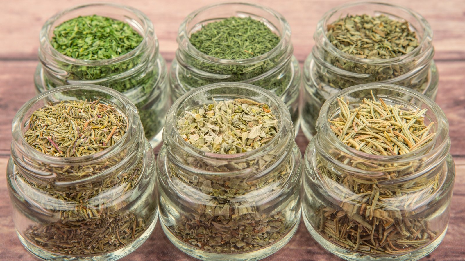 Mason jars filled with dried parsley, tarragon, marjoram, dill weed, thyme, rosemary, and basil sit arranged on a rustic wooden background, showcasing varied green tones and fine, textured leaves.
