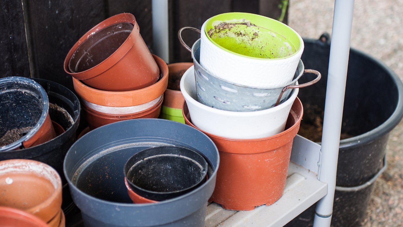 A collection of stacked empty pots in various colors, from terracotta to plastic, rests against a wall, with a layer of dirt and small green tools nearby, suggesting recent use.