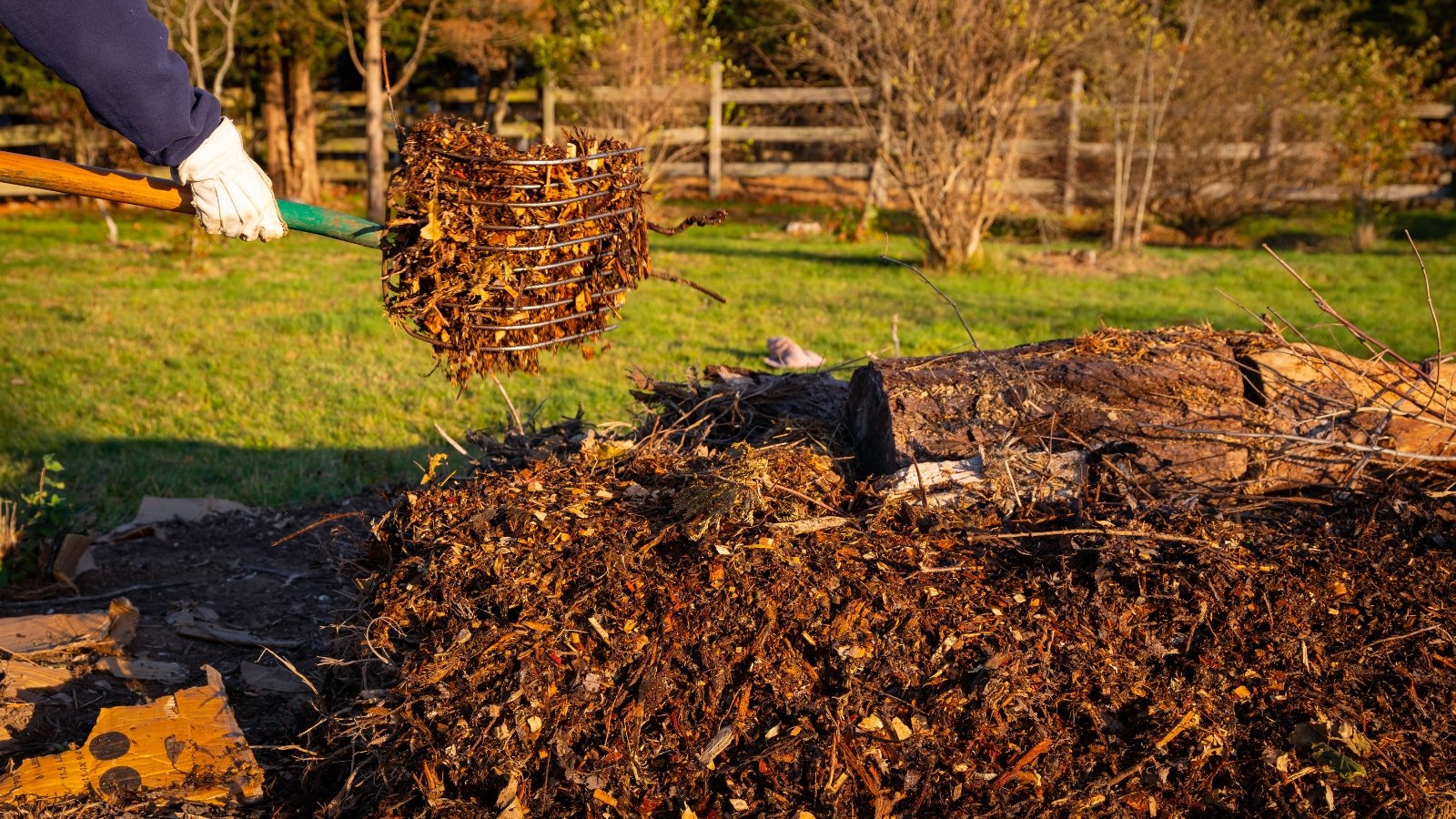 Close-up of a gardener wearing white gloves, using a large garden rake to turn over a pile of orange-brown dry leaves in the garden.