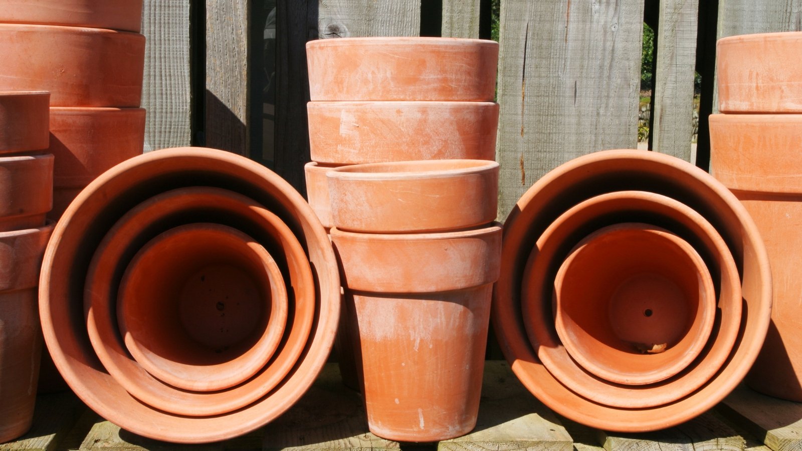 A cluster of empty terracotta clay pots rests beside a wooden fence in the sunny garden.
